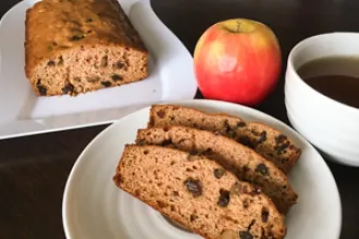 A plate with three slices of applesauce loaf cake in the foreground. In the background, a partial loaf of cake rests on a serving plate next to an apple and cup or bowl of liquid.