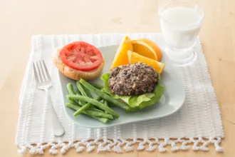 Black bean burgers - USDA Plate with a black bean burger on a bun with green beans and orange wedges. The plates rests on a cloth placemat with a fork and glass of milk next to it.